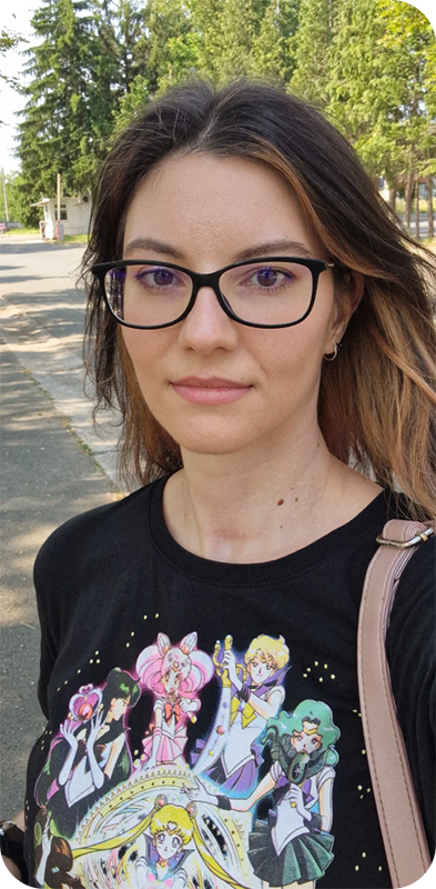 The photograph shows the artist looking at the camera in an outdoor enviroment. The artist is a white woman, in her 30s, with long swept back dark hair, dark eyebrows and eyes. The aritist is wearing glasses, a black T-shirt with an illustration featuring Sailor Moon, and a pink backpack. A road with some trees is visible in the background